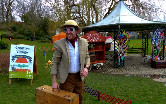 Performer smiling with suitcase stood by gazebo with sign saying "Creative Village".