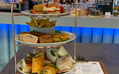 Afternoon tea arranged on table next to menu inside Lime Bar Lounge with bar in background.