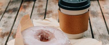 Takeaway coffee cup on a table next to glazed doughnut.